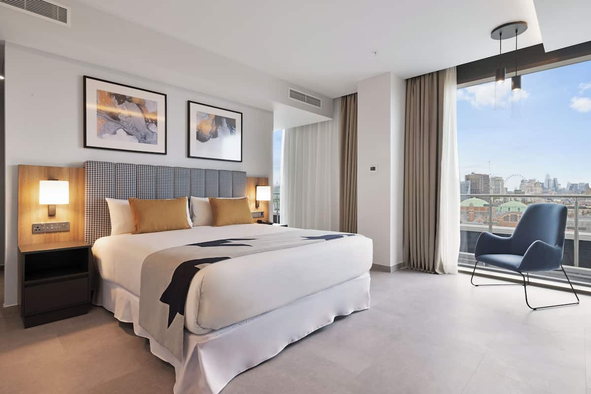 A modern hotel room with a large bed, two bedside lamps, a view of the city, and a balcony chair.