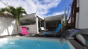 A swimming pool with a water feature, surrounded by a white building with a covered patio area, a palm tree, and a pink chair. - KS VILLA (Saint-François)