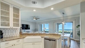 A modern kitchen with white cabinetry, a dishwasher, and a granite countertop. There is a dining area with a table and chairs, and a view of the ocean through large windows.