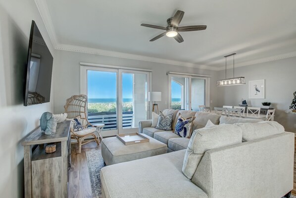 Ein geräumiges Wohnzimmer mit einem großen Sofa, einem Couchtisch und einem Essbereich mit Tisch und Stühlen. Ein Deckenventilator, ein an der Wand montierter Fernseher und ein Blick ins Freie durch große Fenster sind ebenfalls vorhanden.