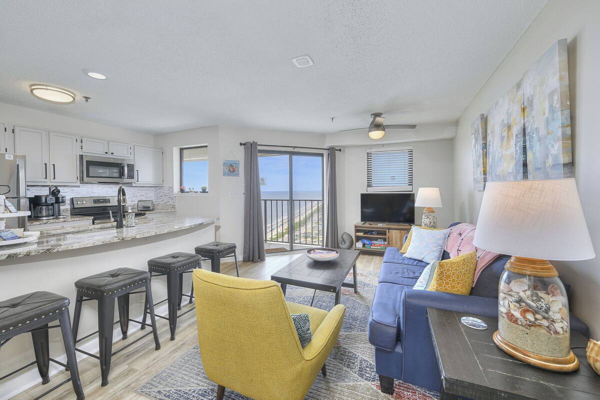 A modern living room with a sofa, a coffee table, and a TV. There is a kitchen area with a microwave and a dining table.