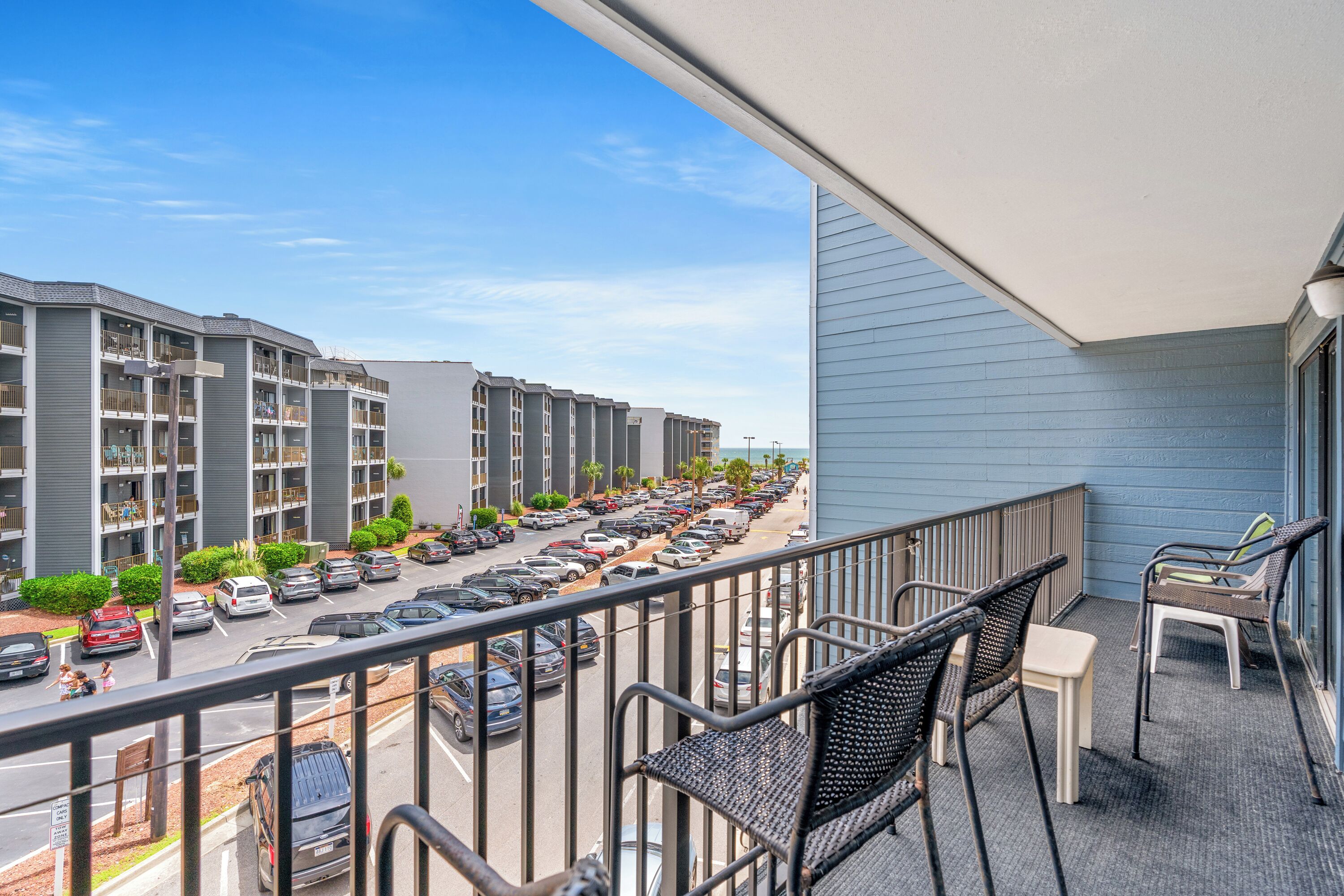 A balcony with outdoor furniture overlooking a parking lot and a row of multi-story buildings.