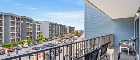 A balcony with outdoor furniture overlooking a parking lot and a row of multi-story buildings.