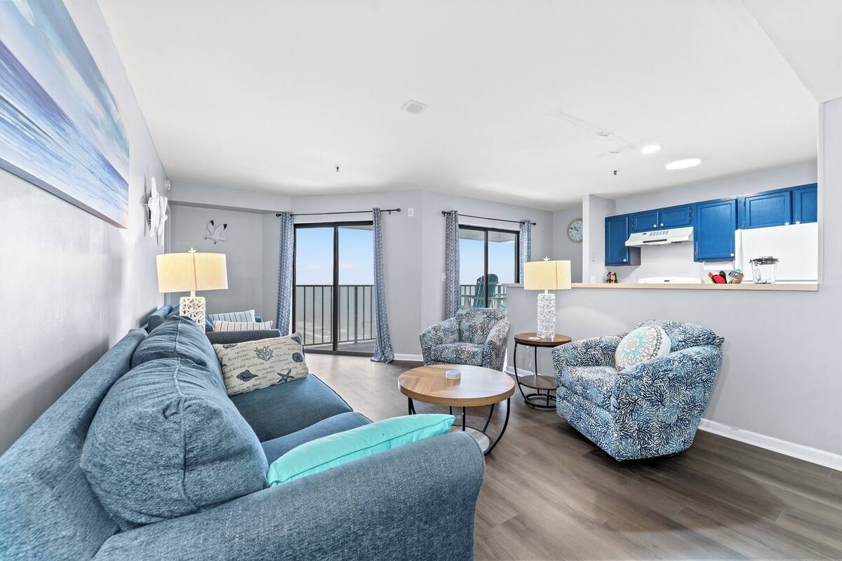 A modern living room with a sofa, armchairs, a coffee table, and a kitchen area with blue cabinets.