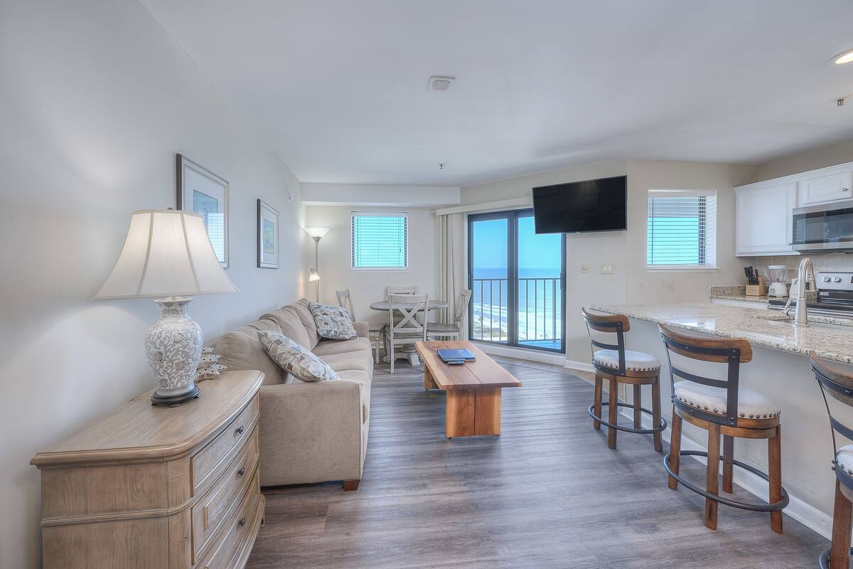 A modern living room with a sofa, coffee table, and chairs, a dining area with a table and chairs, and a kitchen with a TV and counter.