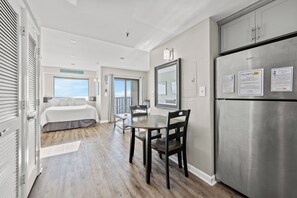 A modern hotel room with a bed, a dining area with a table and chairs, a large window with a view, a refrigerator, and a door leading to a balcony. - Myrtle Beach Resort by Elliott Realty (Myrtle Beach)