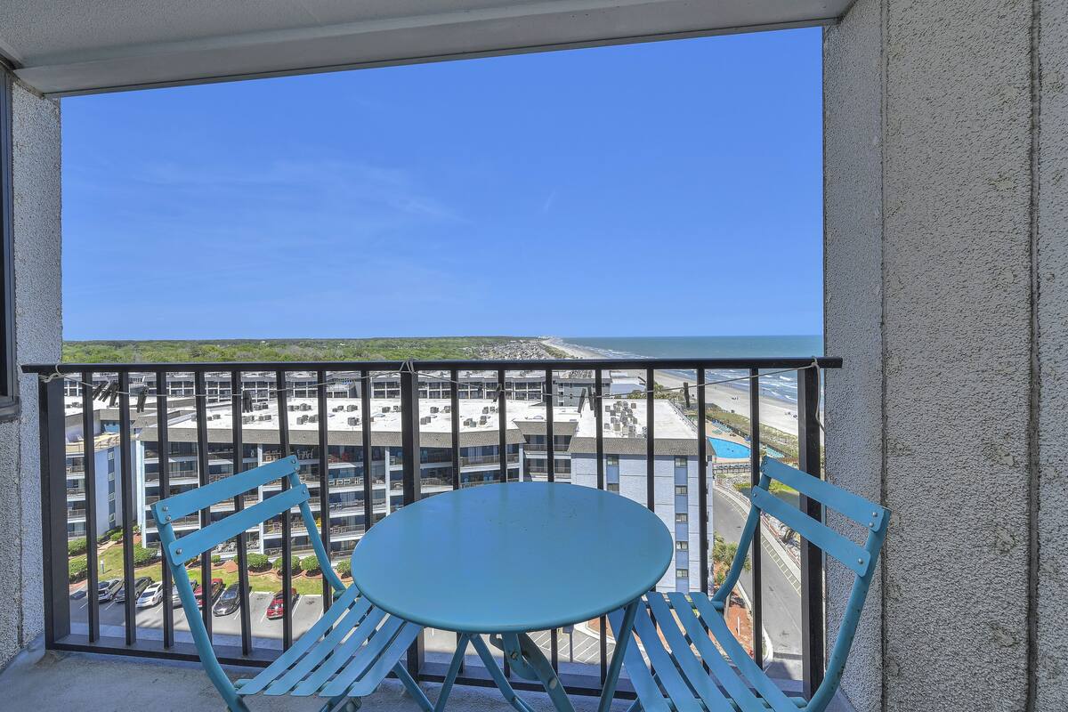 A balcony with a blue table and chairs, overlooking a parking lot and buildings.