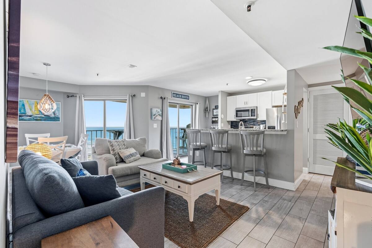 A modern living room with a sofa, coffee table, and artwork; a kitchen with bar stools; and a balcony with a view.