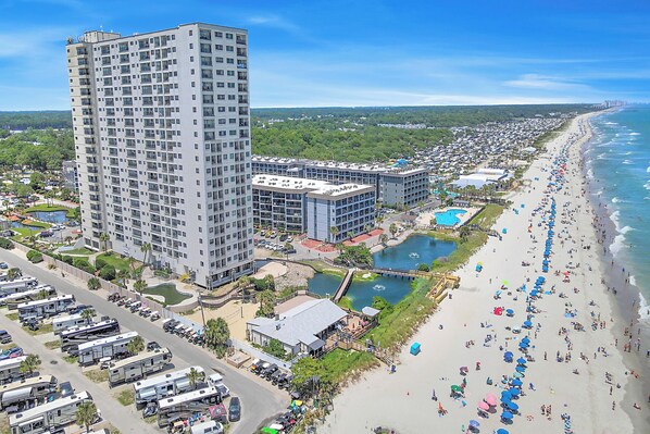 A high-rise hotel with multiple floors, a beach with umbrellas, and a parking lot with RVs. - Myrtle Beach Resort by Elliott Realty (Myrtle Beach)