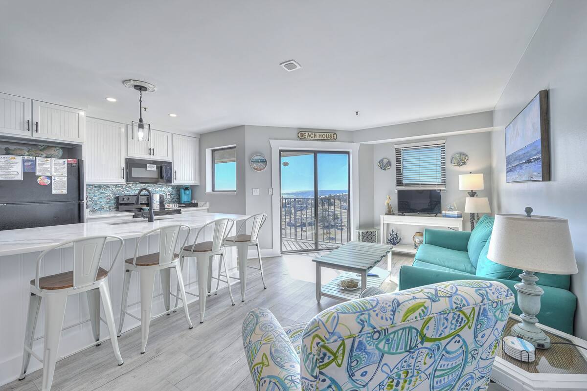A modern kitchen with white cabinets, a microwave, and a dining area with white chairs. There's a teal sofa and a lamp in the living area.