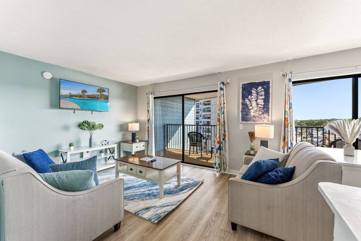 A living room with a sofa, coffee table, and TV, a balcony with a view, and a sliding glass door.