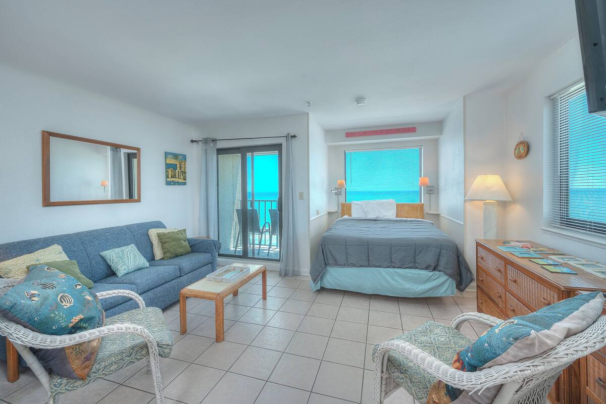 A room with a bed, a sofa, a dining table, and a chair. There is a window with blinds and a view of the outside.