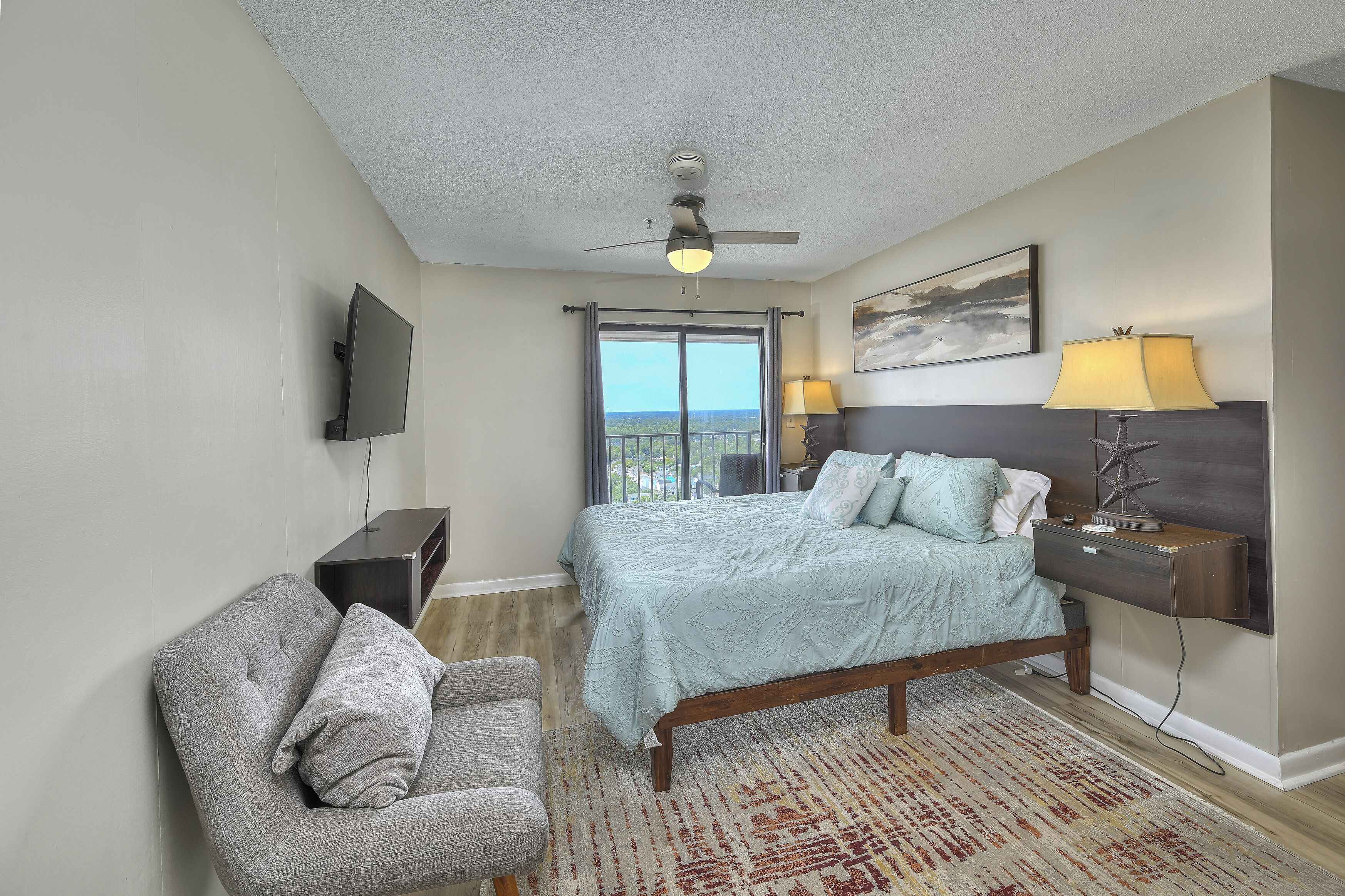 A hotel room with a bed, a desk, a chair, a TV, a ceiling fan, a window with a view, and a lamp.