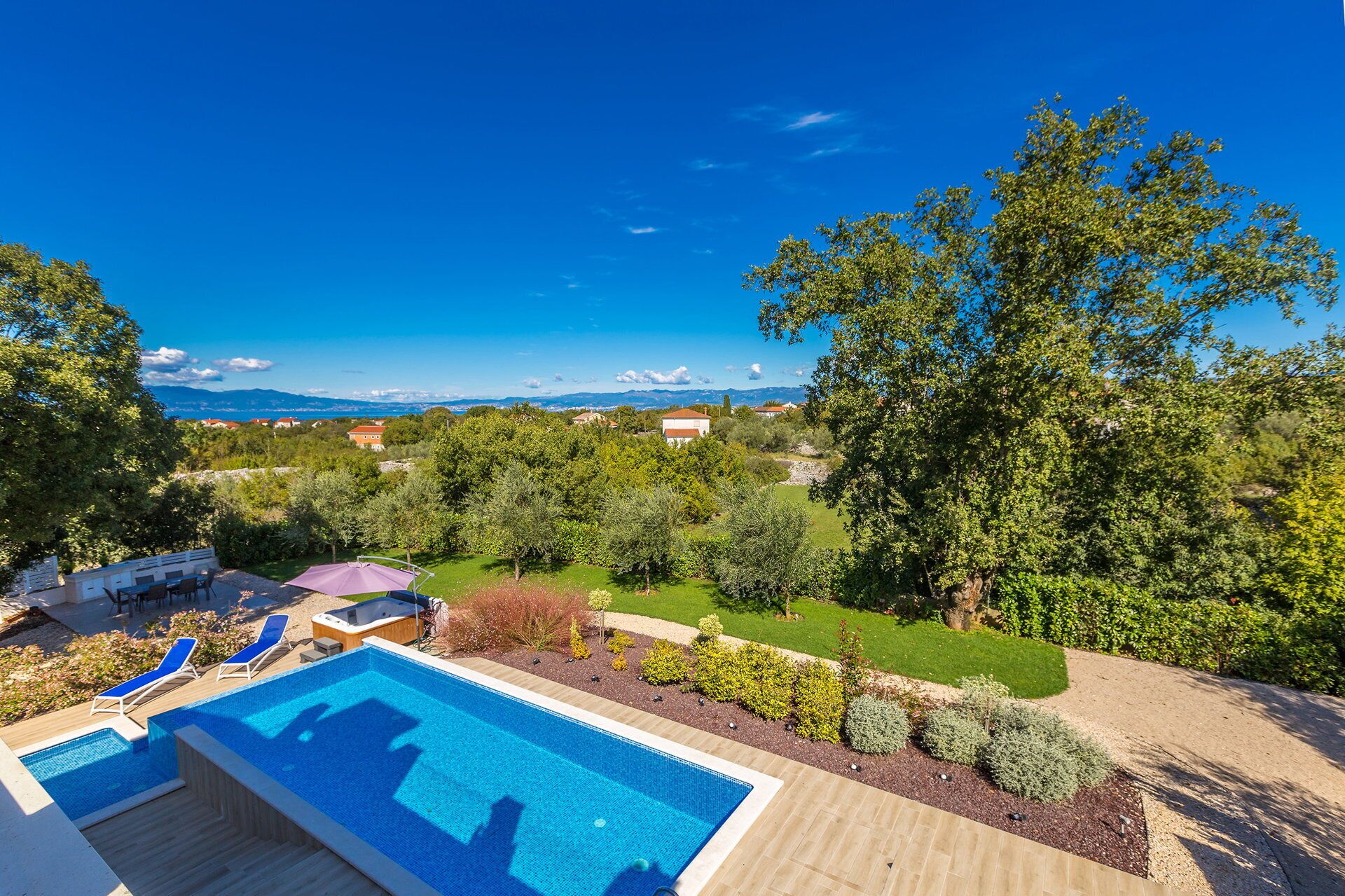 Une piscine entourée de chaises longues, elle-même bordée d’un jardin bien entretenu, d’où l’on peut admirer des maisons et des montagnes au loin.
