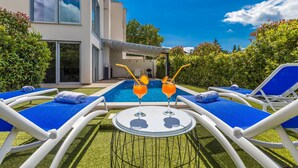 A modern outdoor pool area with lounge chairs and a table, surrounded by greenery and a building with large windows.