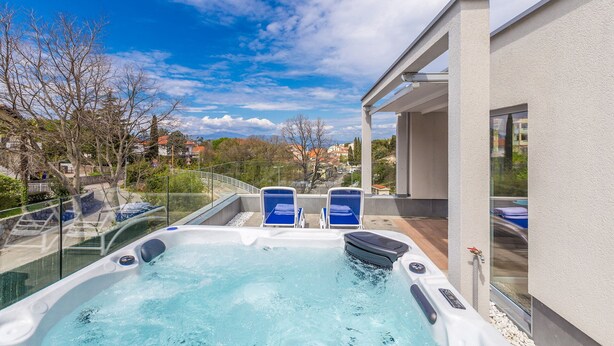 Un jacuzzi con dos sillas azules, una barandilla de cristal y vistas a una zona residencial.