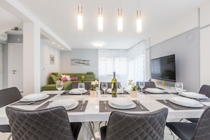 Un espace de salle à manger moderne, avec une longue table dressée pour six personnes, entourée de chaises noires à dossier matelassé. On aperçoit un canapé vert au fond, un téléviseur à écran plat fixé au mur et un tableau au-dessus du canapé.
