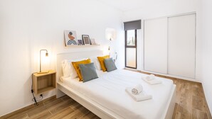 Un dormitorio moderno con una cama, mesitas de noche, un estante en la pared con cuadros, una ventana y un armario.