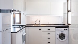 Una cocina compacta con armarios blancos, un lavamanos de acero inoxidable, lavadora integrada y un refrigerador.