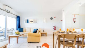 A modern living room with a sofa, a dining table with chairs, a small side table, and a window with blue curtains. - #088 T2 São Rafael 64B by Home Holidays (Albufeira)