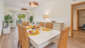 Un coin repas avec une table blanche, des chaises en osier et une table dressée pour le petit-déjeuner, comprenant des fruits et des viennoiseries.