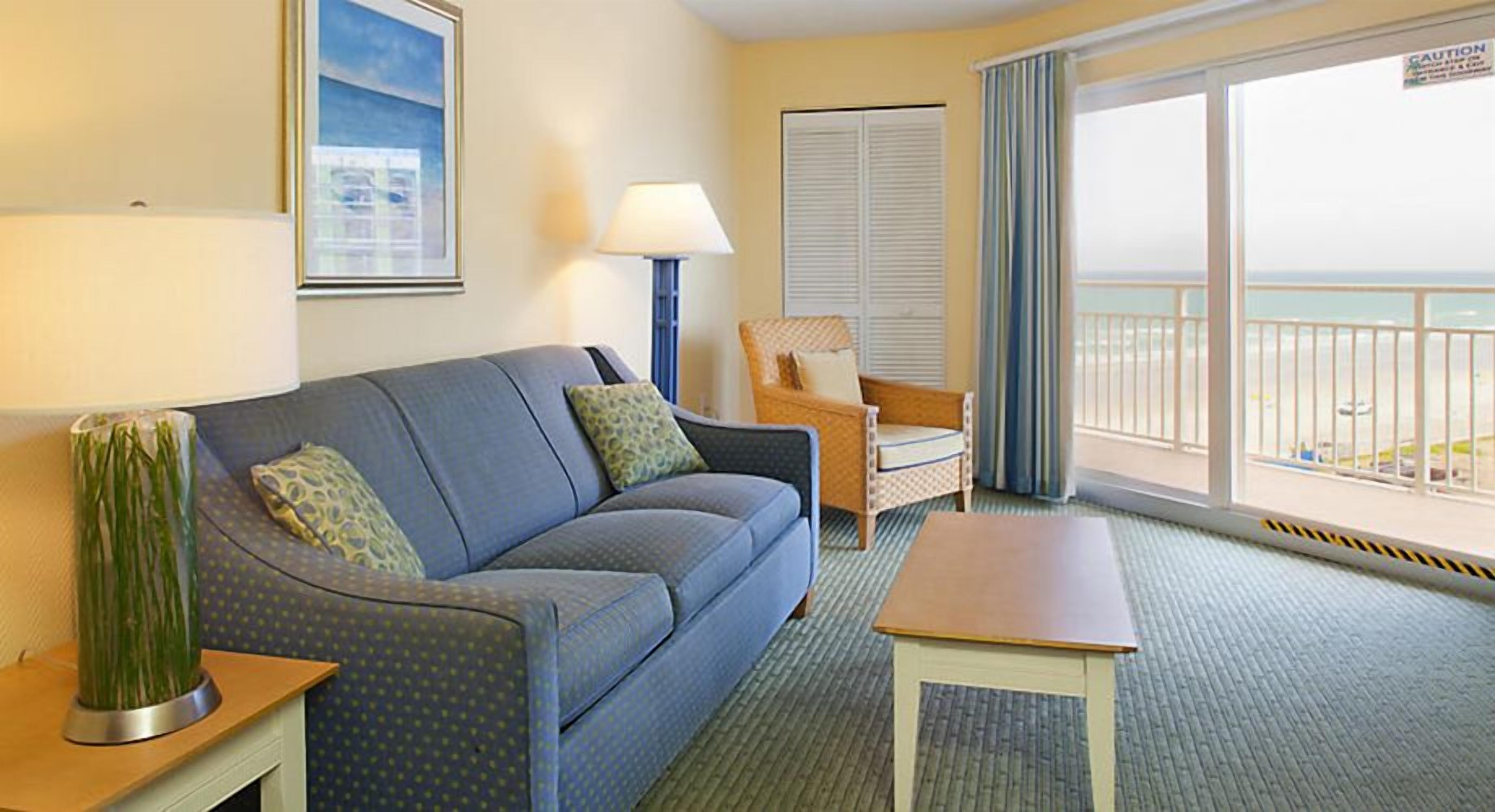 Habitación de hotel con sofá azul, mesa de madera, silla de mimbre, lámpara y ventana grande con vista a la playa.