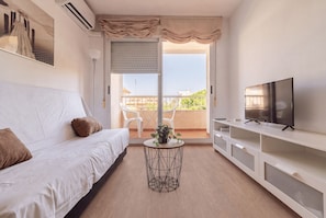 Een moderne woonkamer met een witte bank, een televisie op een witte kast, een ronde salontafel met een plant en een balkon met uitzicht.