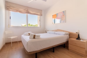 Een slaapkamer met een bed, nachtkastje, lamp, stoel en een raam met uitzicht op gebouwen.