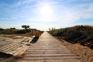A wooden boardwalk leading to a sandy area with palm trees and a clear sky.