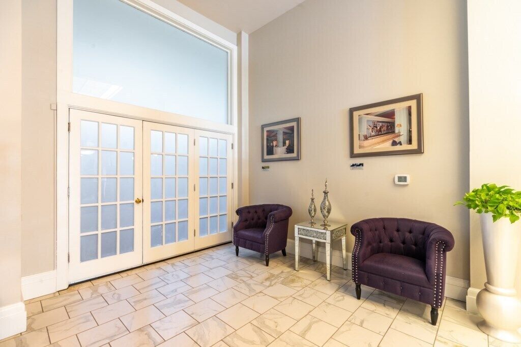 A spacious room with a large sliding glass door leading to a balcony, two armchairs, a small table with decorative items, and framed pictures on the wall.