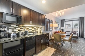 A modern kitchen with dark wood cabinets, a black countertop, and a built-in microwave. There is a dining area with a table and chairs, and a large window with curtains.