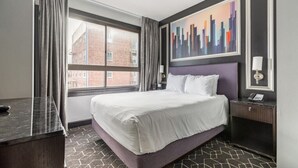 A hotel room with a large bed, a window with curtains, a cityscape painting, and a bedside table with a lamp.