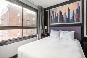 A hotel room with a large bed, a cityscape painting, a window with curtains, and a view of a brick building.