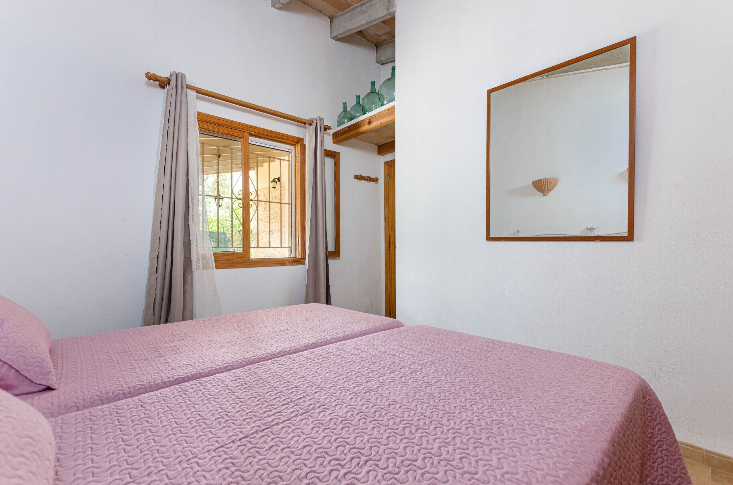 A bedroom with a bed, a window with curtains, a mirror, and a shelf with green bottles.