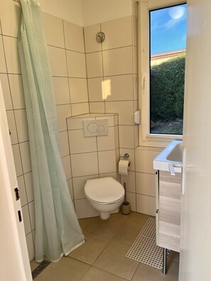 Ein kleines, sauberes Badezimmer mit Toilette, Duschvorhang, Fenster und Waschbecken.