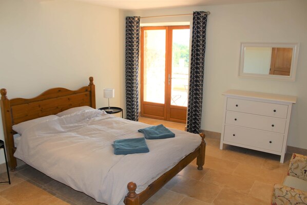 Une chambre avec un lit en bois, une table de chevet blanche, une commode blanche et une porte donnant sur un balcon.