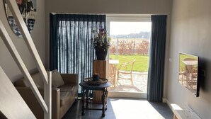 A spacious living room with a sofa, a coffee table, and a TV mounted on the wall. A staircase leads to an upper level, and a large window offers a view of the outdoors. - Group accommodation 32 pers, near Efteling and Beekse Bergen (Haghorst)