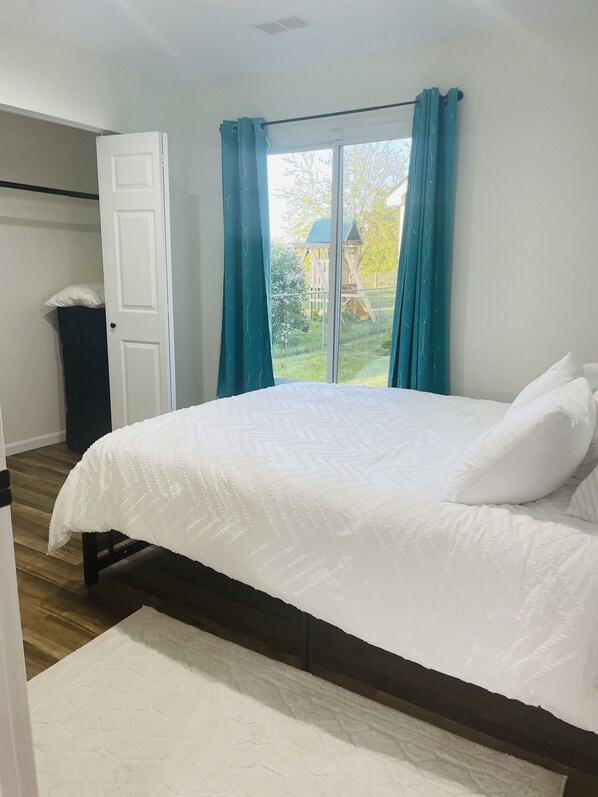 Un dormitorio con cama, ventana con cortinas, vista a un parque infantil y una puerta.