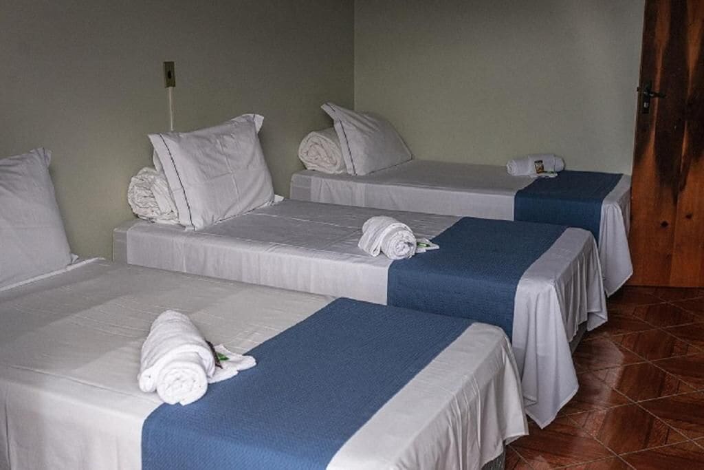Quarto de hotel com três camas de solteiro, cada uma com colchão branco e colcha azul. Há almofadas brancas em cada cama, e toalhas dobradas cuidadosamente sobre as camas. O quarto tem uma porta de madeira e piso de azulejos.