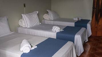 Quarto de hotel com três camas de solteiro, cada uma com colchão branco e colcha azul. Há almofadas brancas em cada cama, e toalhas dobradas cuidadosamente sobre as camas. O quarto tem uma porta de madeira e piso de azulejos.