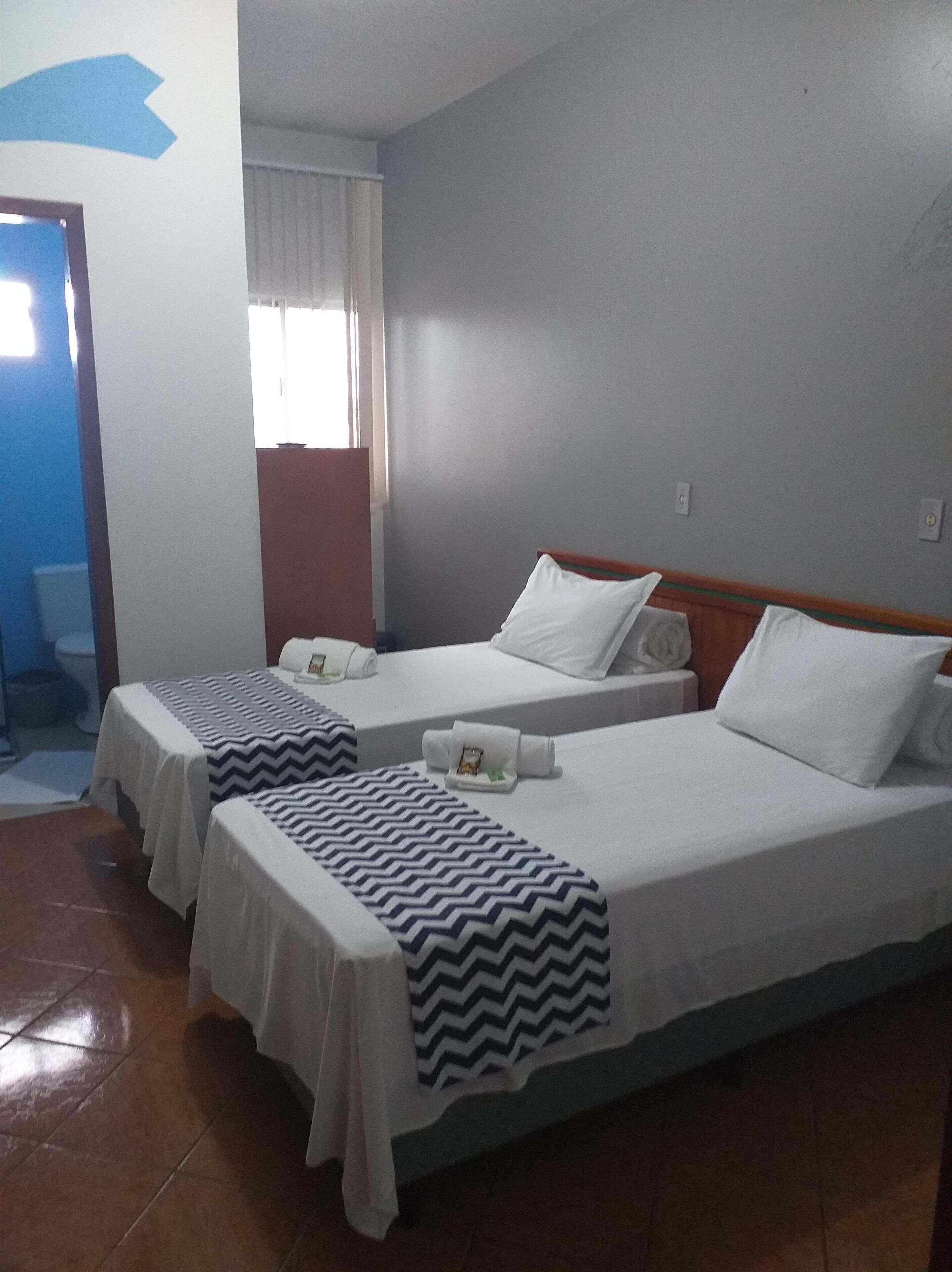 A hotel room with two beds, each with white linens and a zigzag patterned blanket. There is a window with white curtains, a wooden door, and a tiled floor.