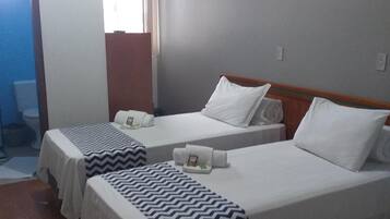 A hotel room with two beds, each with white linens and a zigzag patterned blanket. There is a window with white curtains, a wooden door, and a tiled floor.