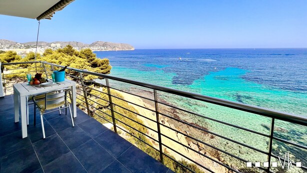 A balcony with a table set for two, overlooking a coastal area with clear turquoise waters and distant hills. - Maite Apartment in Moraira – Seafront holiday apartment for 4 guests with air conditioning and Wi-Fi (Moraira)
