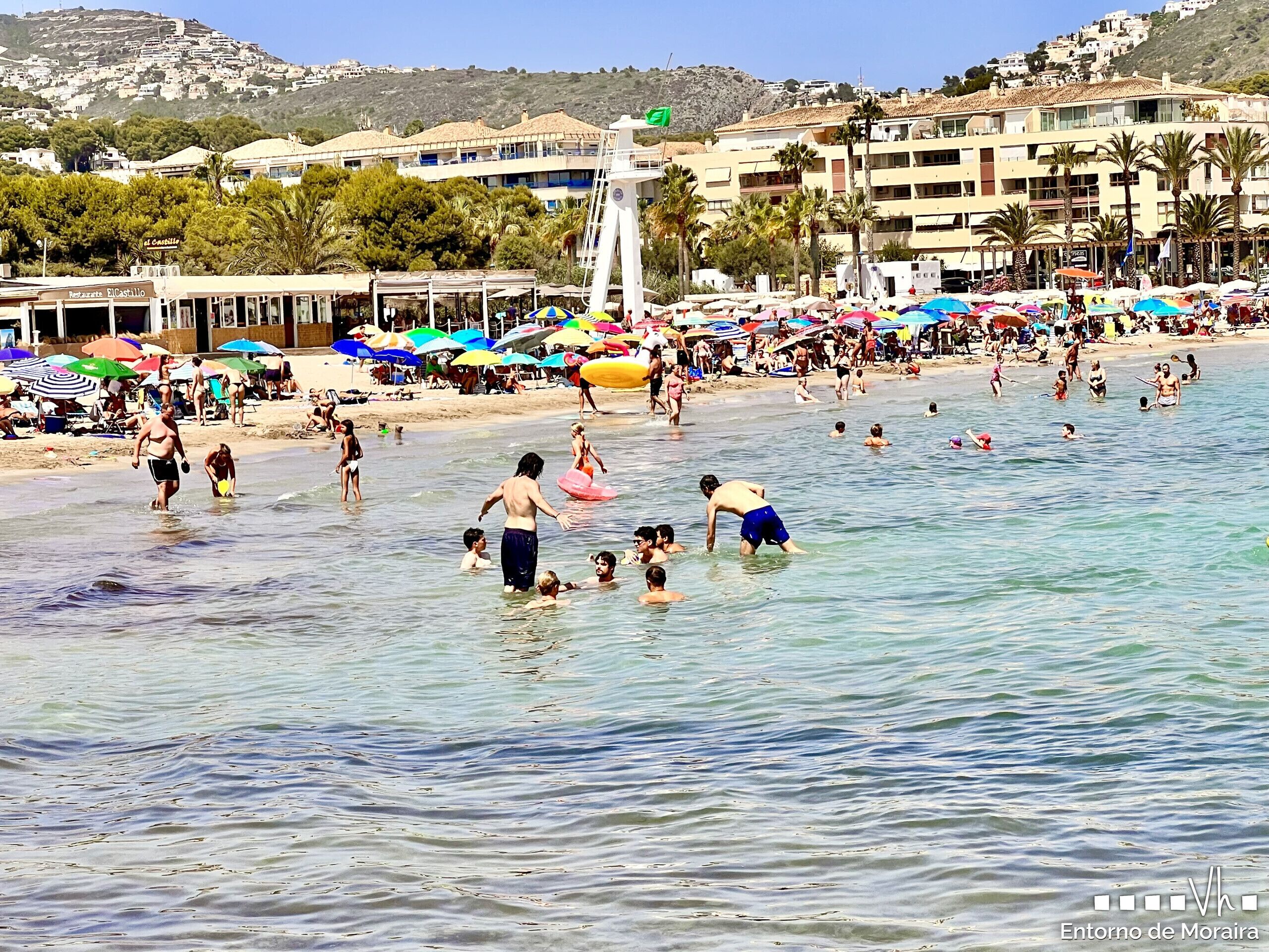 Een druk strand met mensen die zwemmen en zonnebaden, parasols en gebouwen op de achtergrond.