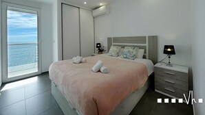 A modern bedroom with a large bed, a nightstand, a lamp, and a view of the sea through a sliding door. - Maite Apartment in Moraira – Seafront holiday apartment for 4 guests with air conditioning and Wi-Fi (Moraira)