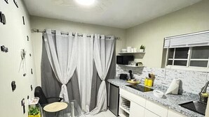 Cozinha compacta com área de refeições, com mesa de vidro, cadeiras pretas, forno embutido e janela com persianas.