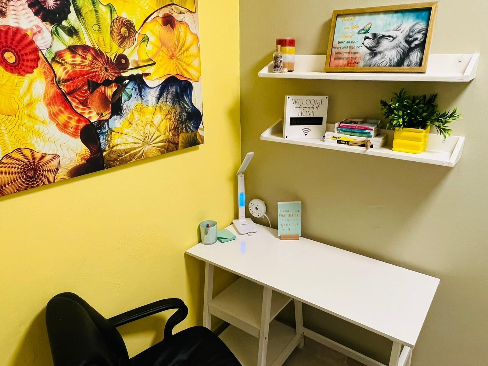 A room with a desk, chair, shelves with books and a framed picture, a clock, and a wall with a colorful abstract painting.