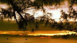 Um pôr do sol sobre um mar calmo, com barcos, uma praia arenosa e um molhe rochoso.