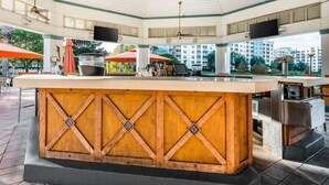 Ein überdachter Außenbereich mit Bar, einem Holzbalken, mehreren Sonnenschirmen und Blick auf Wohnhäuser im Hintergrund.