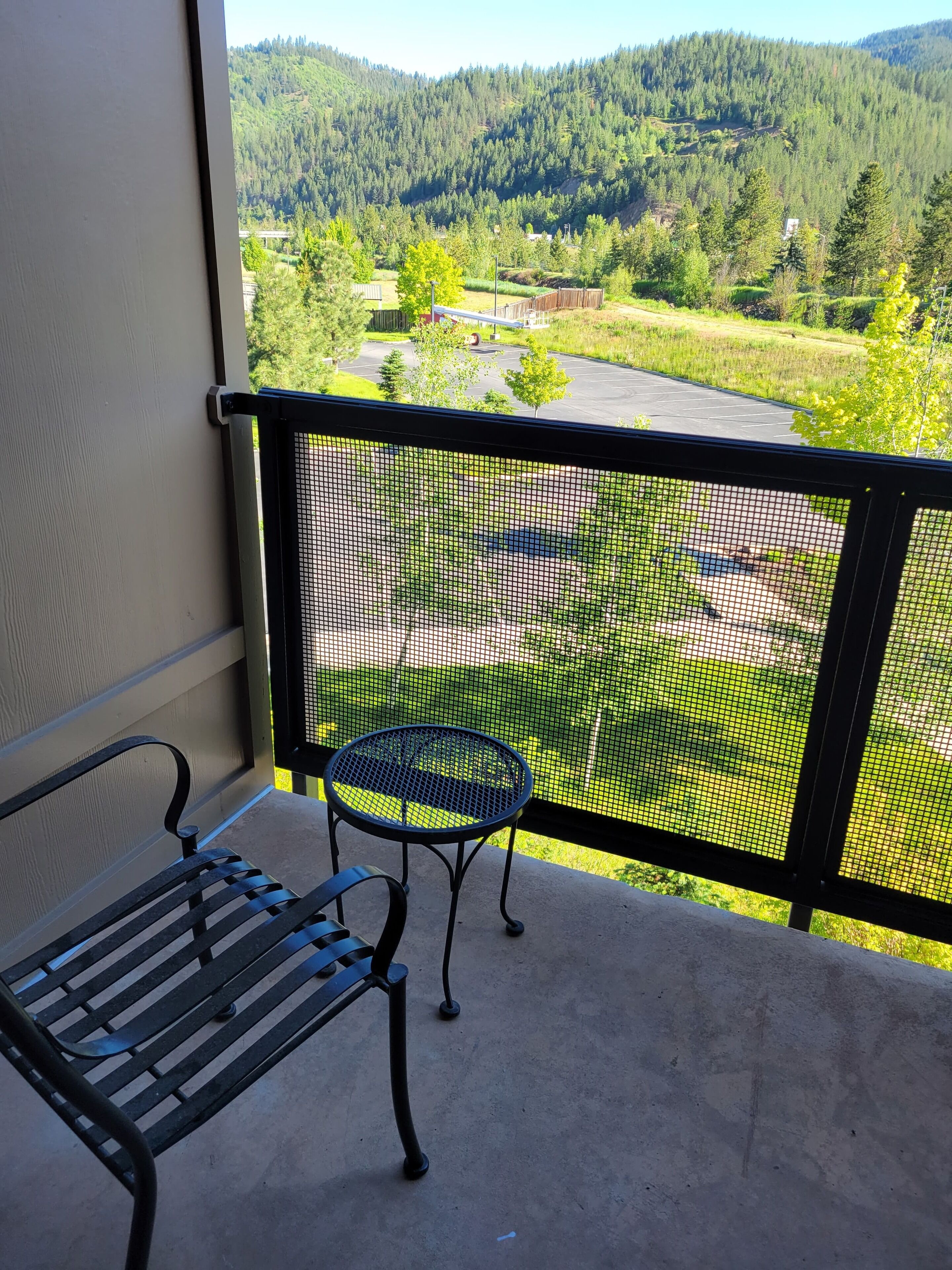 Un balcone con una panca in metallo e un tavolino, affacciato su un paesaggio verde con alberi e un'area parcheggio.